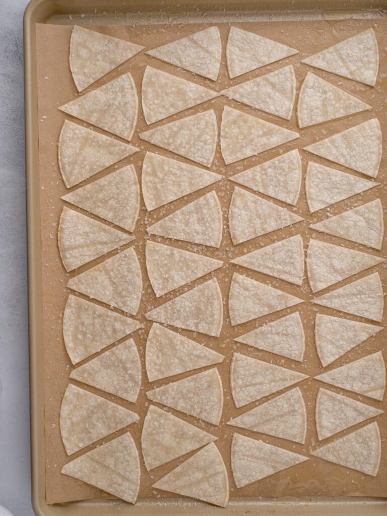 Baking tray with corn tortillas cut into triangle pieces, topped with avocado oil, salted and ready to bake.