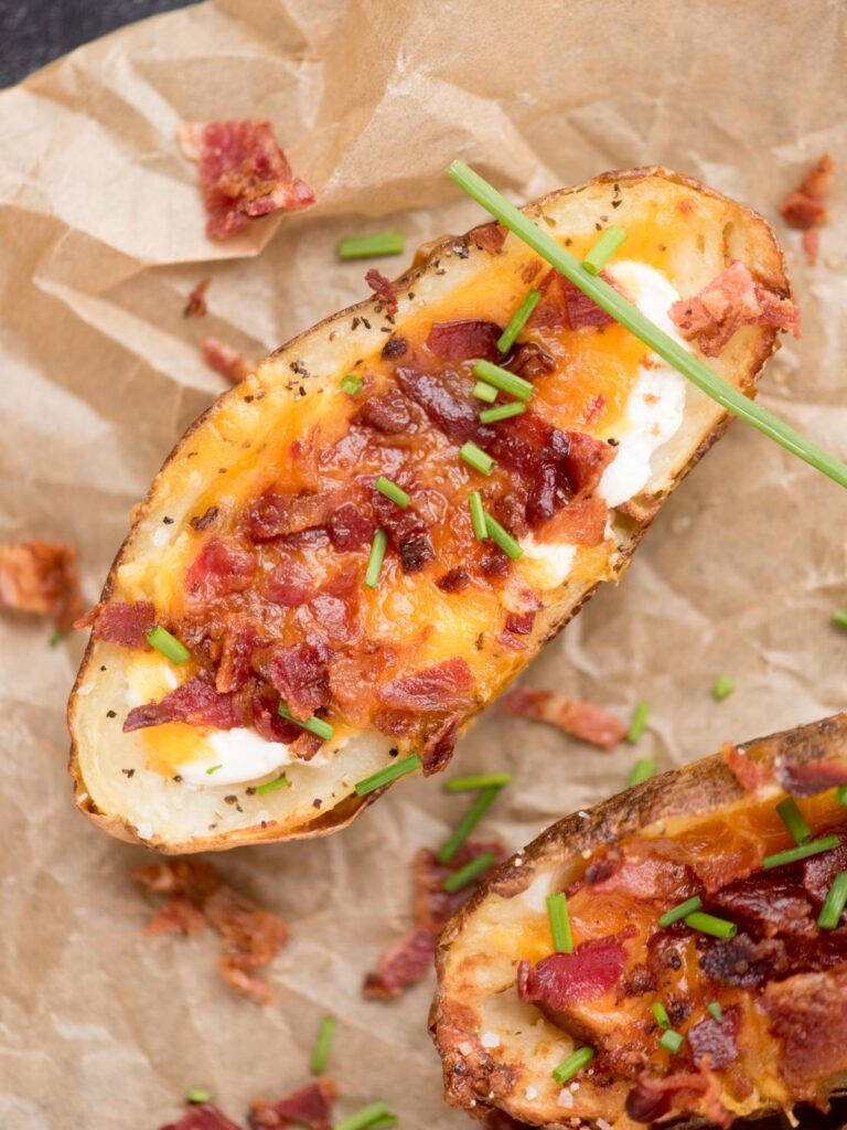 Two air fryer potato skins filled with cheese, bacon and chives on top of parchment paper.