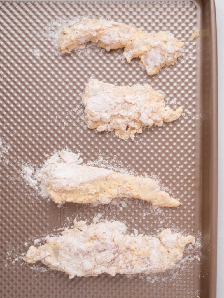 Chicken pieces coated in seasoned flou added to a baking sheet.