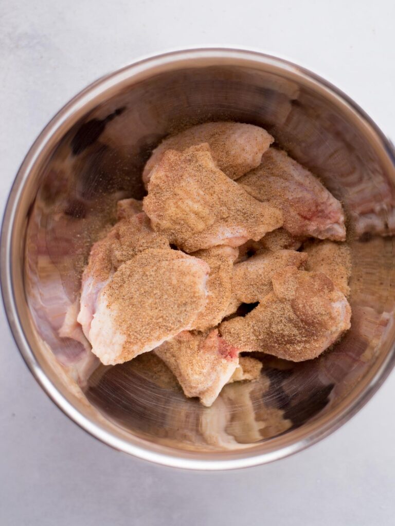 Mixing bowl with raw chicken wings and seasonings sprinkled over the top.