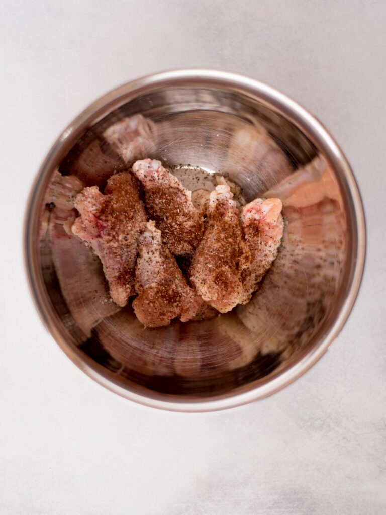 Raw chicken wings with seasonings added over them in a mixing bowl ready to combine.