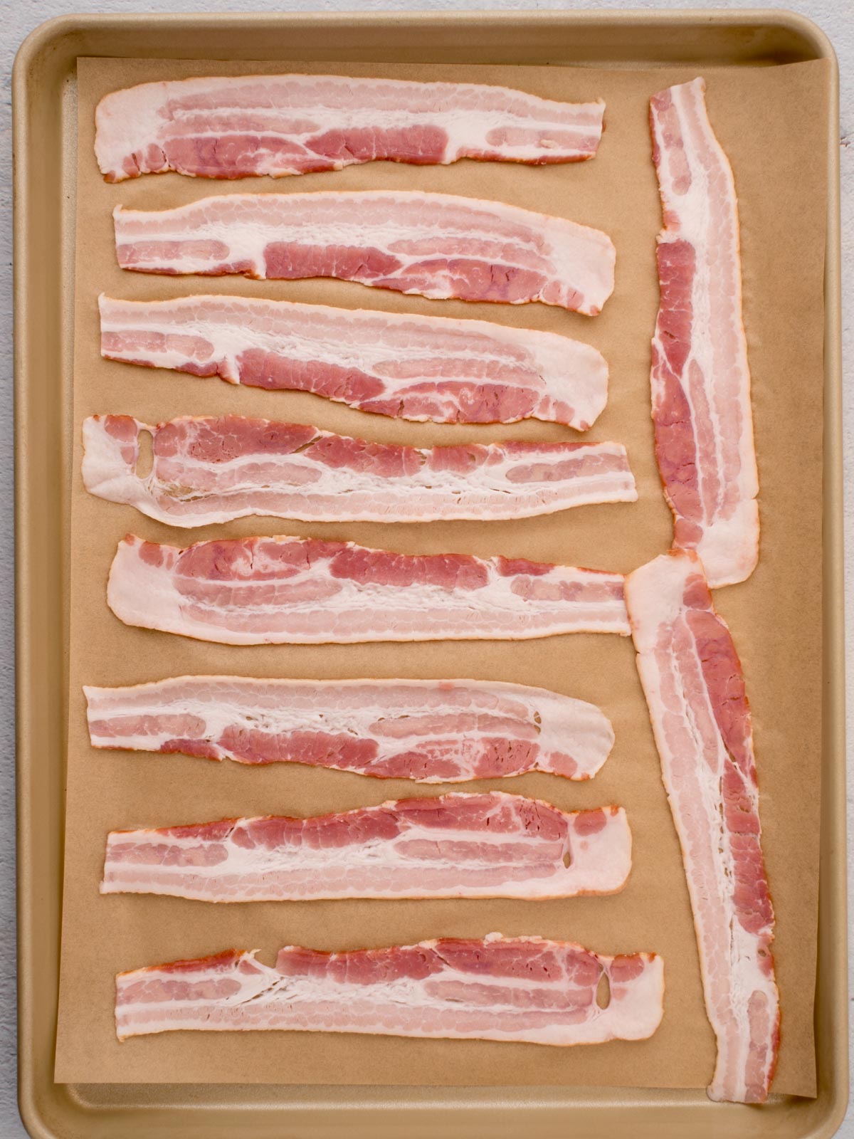 Raw bacon strips over parchment paper on a baking sheet, ready to go into the oven.