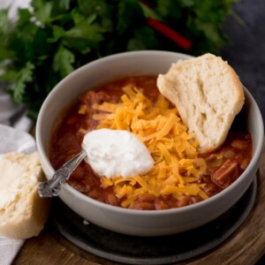 brisket chili in a bowl with a dollop of sour cream, shredded cheese, and half of a buttered roll in the bowl.