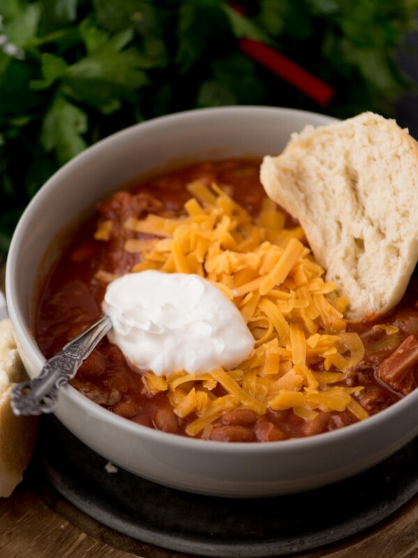 brisket chili in a bowl with a dollop of sour cream, shredded cheese, and half of a buttered roll in the bowl.