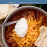A hearty bowl of brisket chili topped with shredded cheese and a dollop of sour cream, served alongside a warm biscuit and a spoon on a rustic wooden surface. Text above reads brisket chili and sweetlysplendid.com.