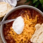 A bowl of brisket chili topped with shredded cheese and sour cream, served with biscuit pieces. A buttered biscuit is on the side. Text above reads award-winning brisket chili. Fresh parsley is also visible, adding a pop of color to this hearty brisket chili dish.