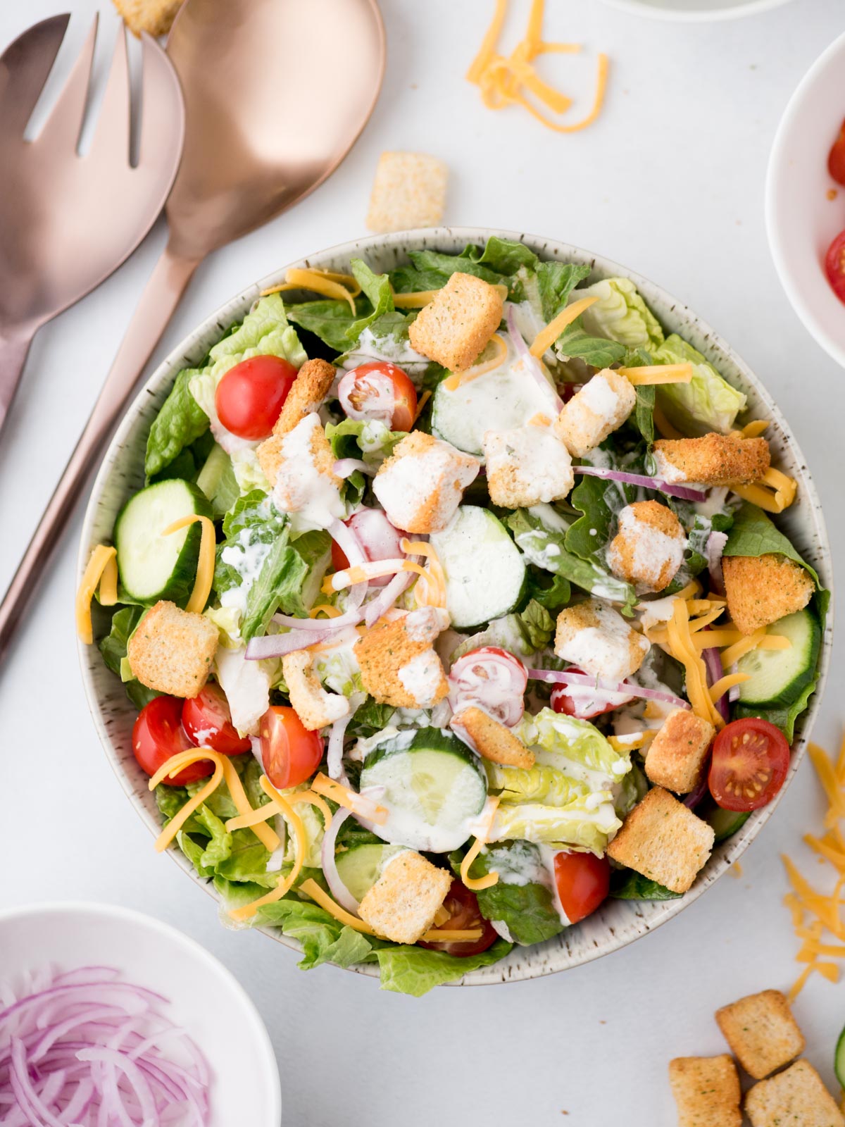 house salad in a bowl topped with ranch dressing, surrounded by servings utensils and toppings