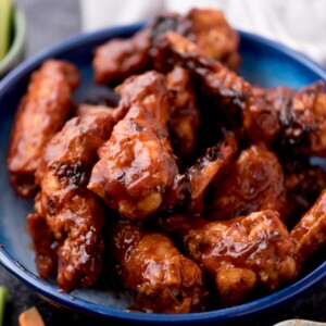 baked bbq chicken wings stacked in a blue bowl surrounded by ranch, fresh celery sticks, and carrot sticks