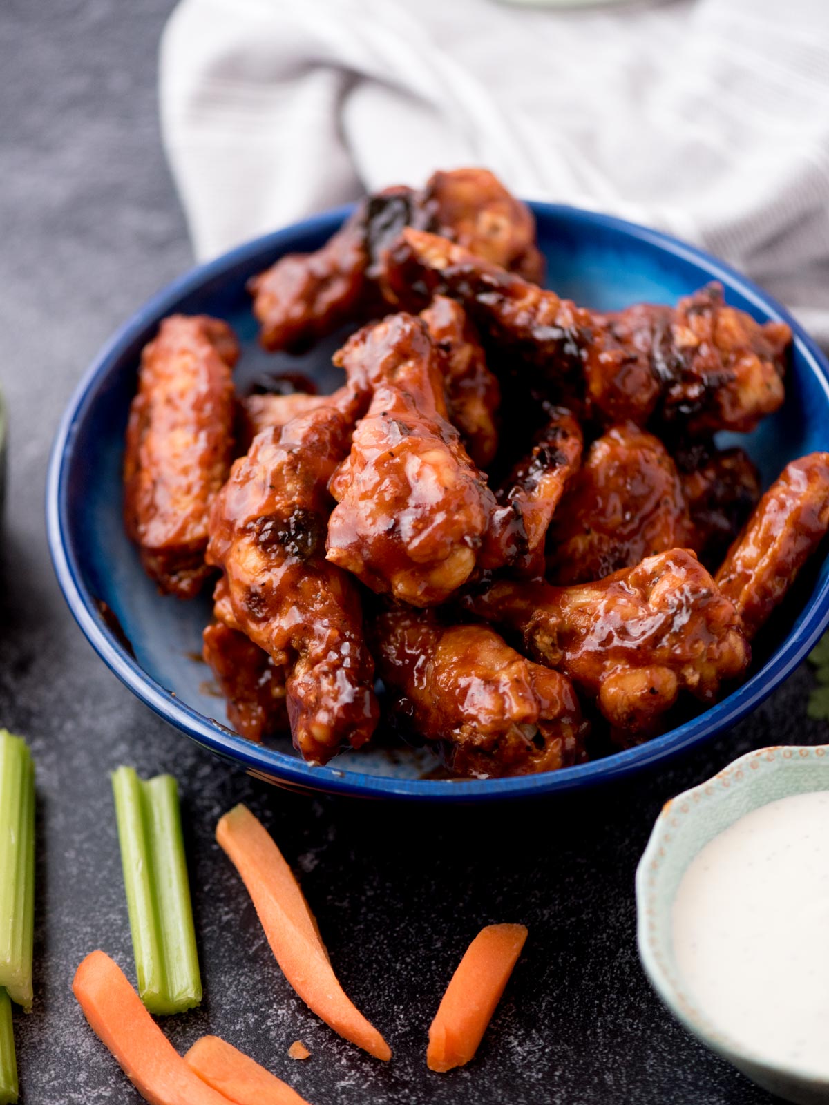 baked bbq chicken wings stacked in a blue bowl surrounded by ranch, fresh celery sticks, and carrot sticks