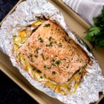 baked salmon in foil on a baking sheet surrounded by fresh parsley and a kitchen towel
