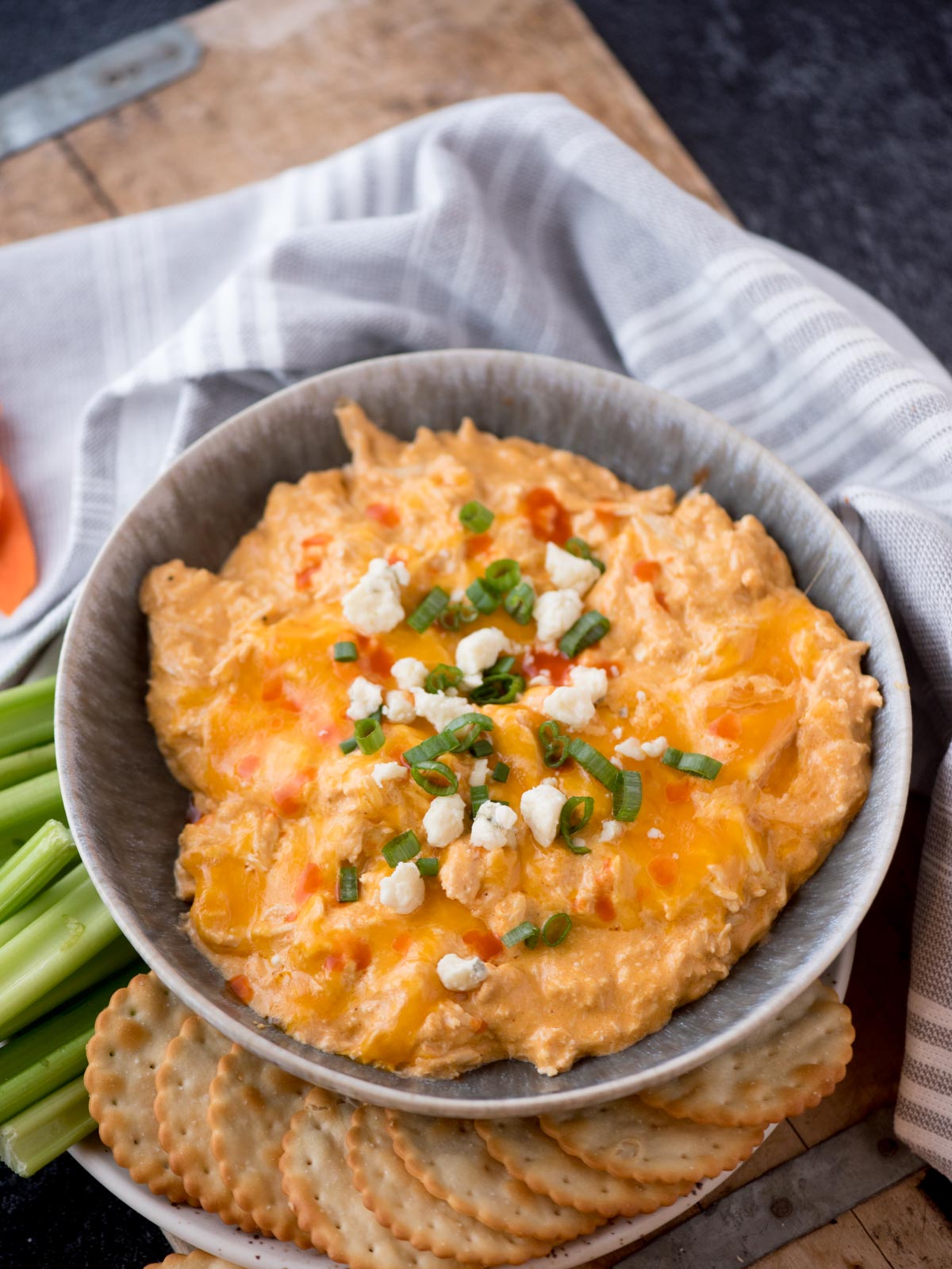 buffalo chicken dip in a serving bowl topped with sliced green onions and blue cheese surrounded by crackers and celery