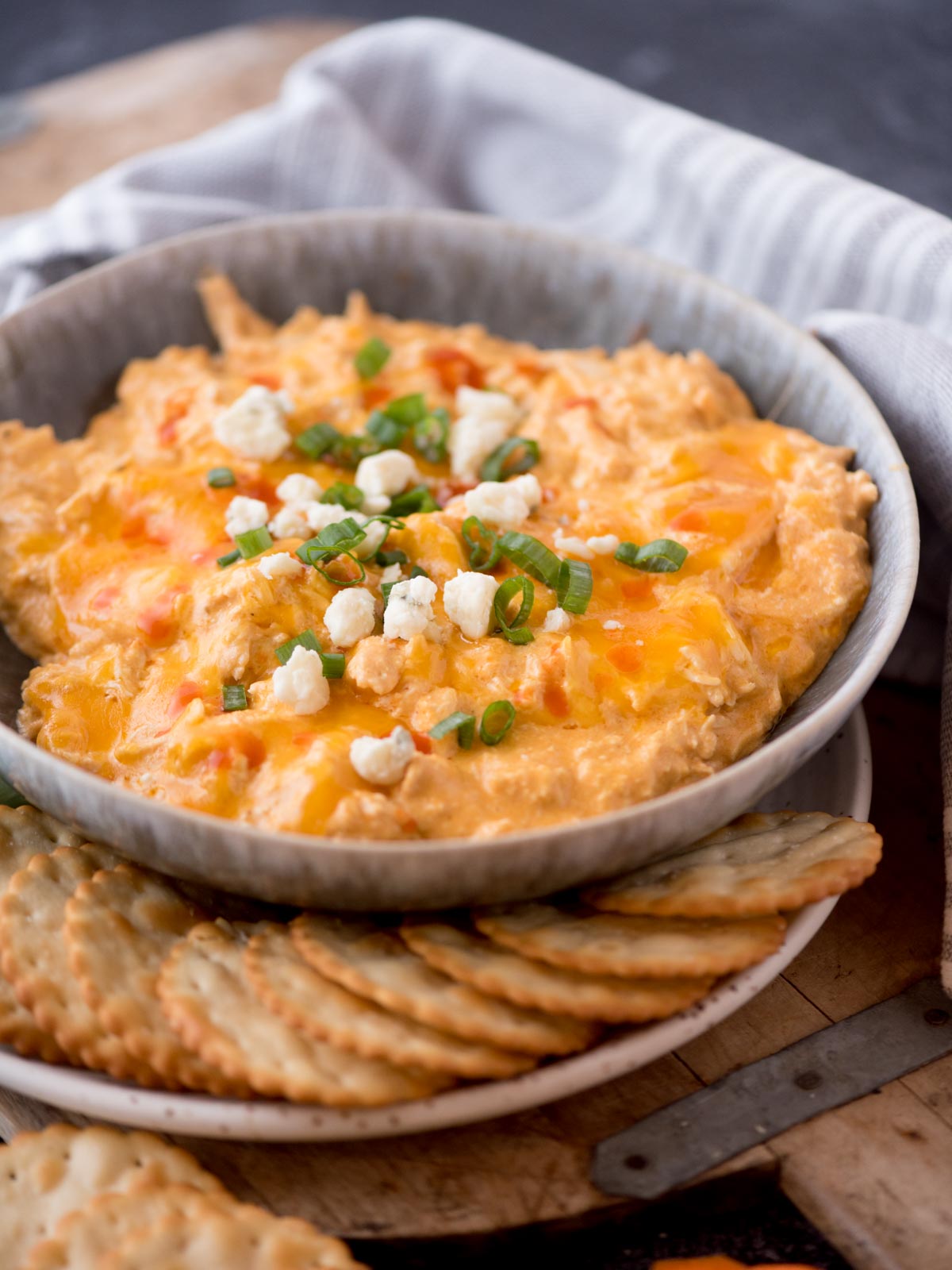 slow cooker buffalo chicken dip in a bowl topped with sliced green onion and blue cheese surrounded by crackers and celery