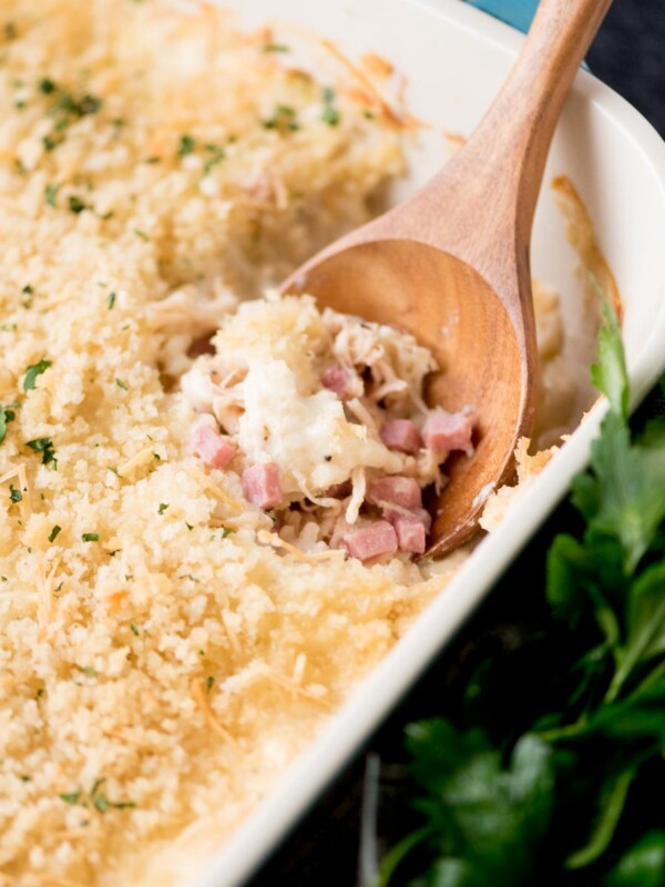 chicken cordon bleu casserole in a baking dish with a serving spoon inside. It is surrounded by fresh parsley and a kitchen towel.