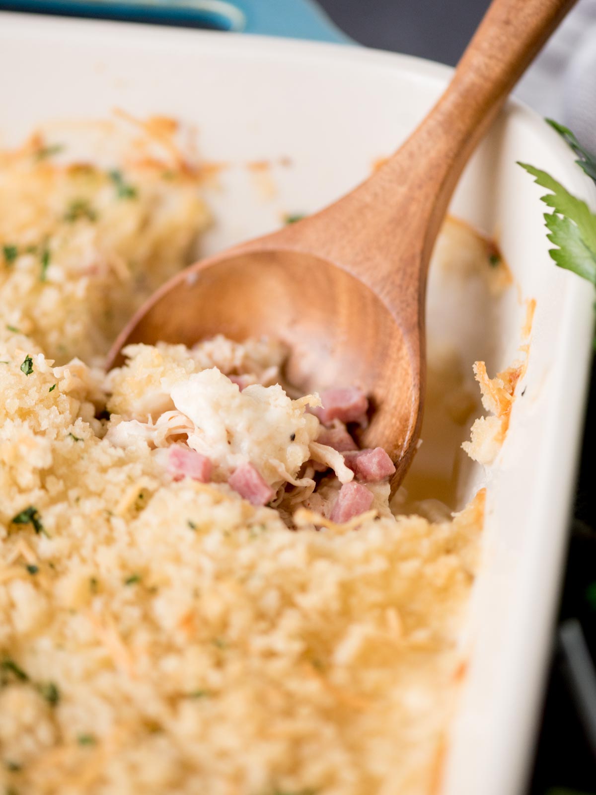 chicken cordon bleu casserole in a baking dish with a serving spoon inside. It is surrounded by fresh parsley and a kitchen towel.