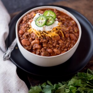 chili in a bowl sitting on a black plate. The chili is topped with shredded cheese, cracked black pepper, sour cream, and jalapeno