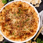 jalapeno popper dip in a white baking dish surrounded by serving utensils, chips, crackers, jalapenos, and parsley