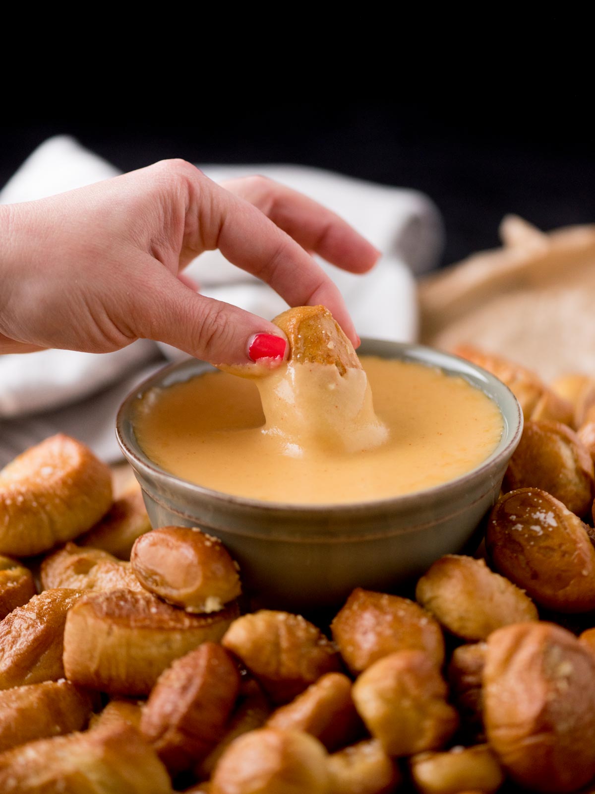pretzel bites being dipped into beer cheese dip in a bowl. It is surrounded by pretzel bites on parchment paper