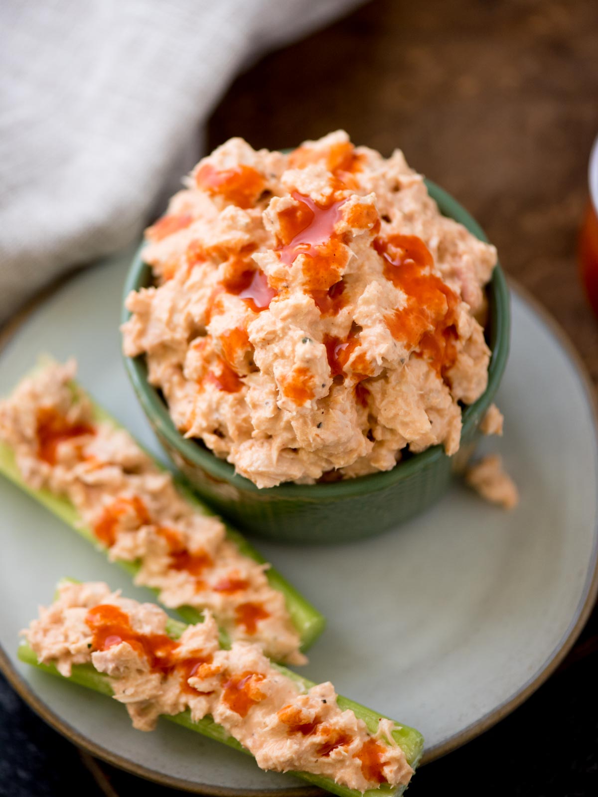 buffalo tuna salad in a bowl with tuna salad stuffed in celery on a plate. It's surrounded by buffalo sauce on a bread board and a kitchen towel.