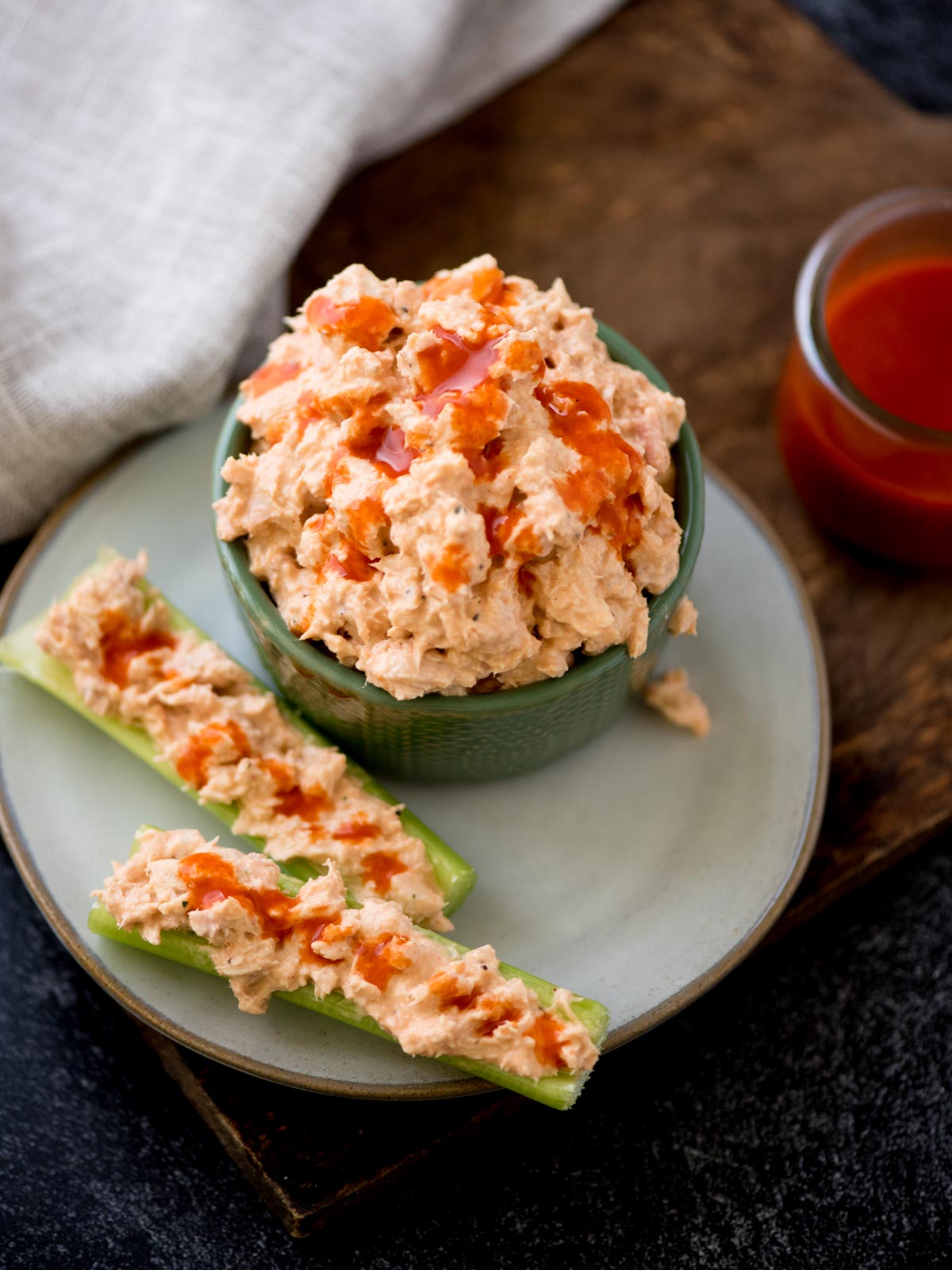 buffalo tuna salad in a bowl with tuna salad stuffed in celery on a plate. It's surrounded by buffalo sauce on a bread board and a kitchen towel.