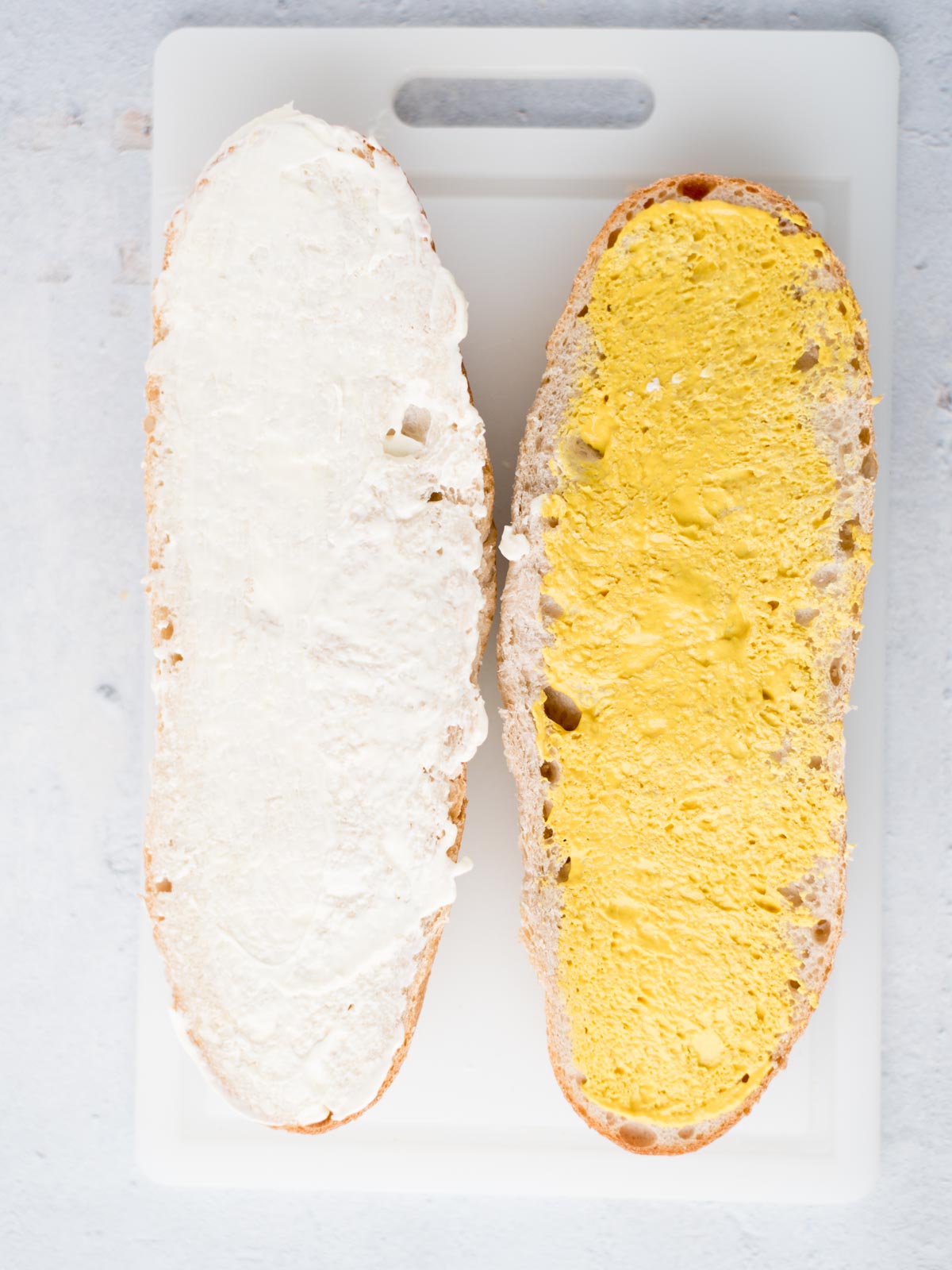 halves of a french bread loaf. One is covered in mayo and the other is covered in mustard.