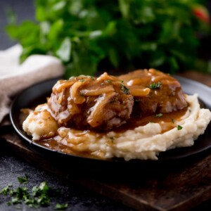 hamburger steak with gravy over a pile of mashed potatoes sitting on a black plate. The plate is on top of a wooden board with parsley in the background.