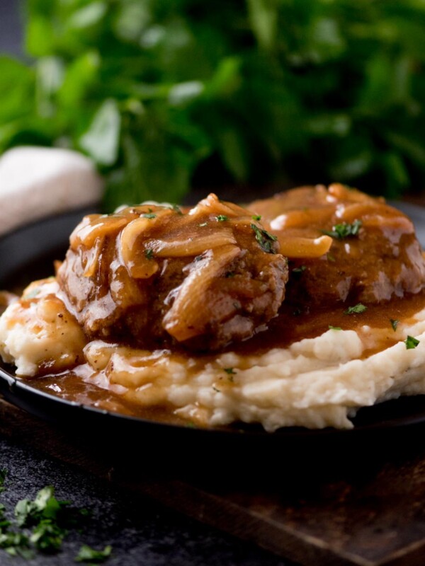 hamburger steak with gravy over a pile of mashed potatoes sitting on a black plate. The plate is on top of a wooden board with parsley in the background.