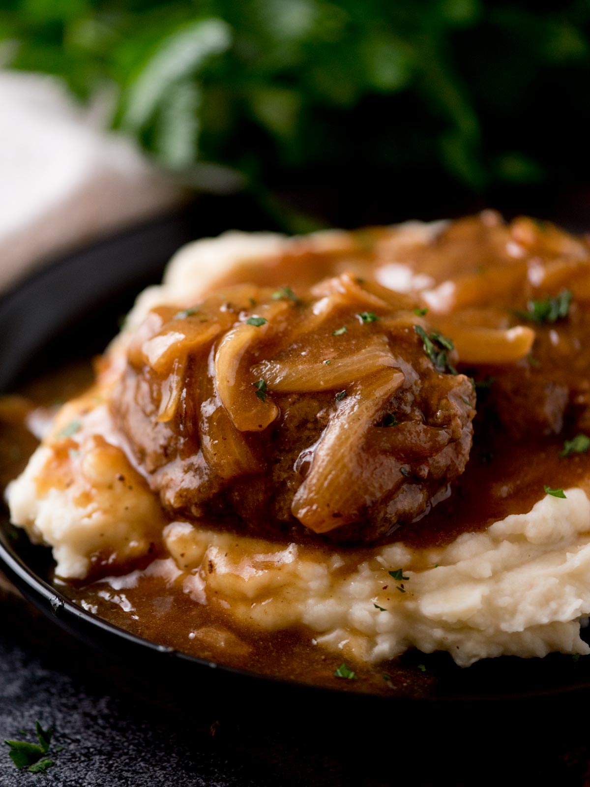 hamburger steak with gravy over a pile of mashed potatoes sitting on a black plate. The plate is on top of a wooden board with parsley in the background.