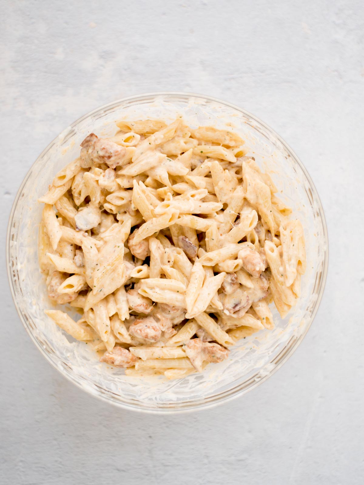 Pasta and cooked chicken pieces coated in white sauce in a mixing bowl.