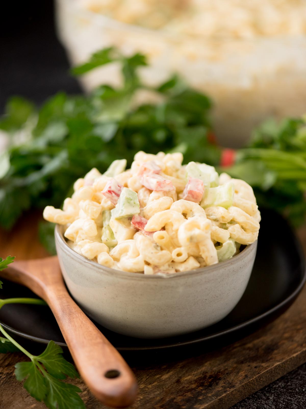 Macaroni salad in a grey bowl surrounded by a serving spoon and fresh parsley.