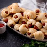 Small bowl of ketchup next to a platter of golden baked pigs in a blanket ready to enjoy.