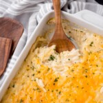 Close-up of a creamy hash brown casserole with a golden, melted cheese topping, being scooped with a wooden spoon from a white baking dish. The casserole is garnished with finely chopped parsley, and the dish is placed on a striped gray and white cloth with wooden utensils beside it.