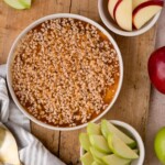 caramel apple dip in a white dish on a wooden board surrounded by slices of apple in bowls, a dish towel, and gold utensils