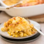A close-up of a serving of corn casserole on a small, round plate with a fork on the side. The casserole appears creamy with bits of whole corn kernels visible. A baking dish with more casserole is blurred in the background.