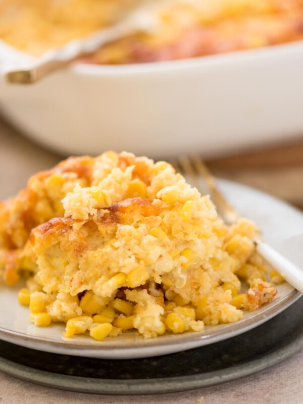 A close-up of a serving of corn casserole on a small, round plate with a fork on the side. The casserole appears creamy with bits of whole corn kernels visible. A baking dish with more casserole is blurred in the background.