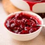 A white bowl filled with vibrant red cranberry sauce, containing whole cranberries and a glossy texture. A spoon is partially submerged in the sauce. The background shows more sauce in a white dish.