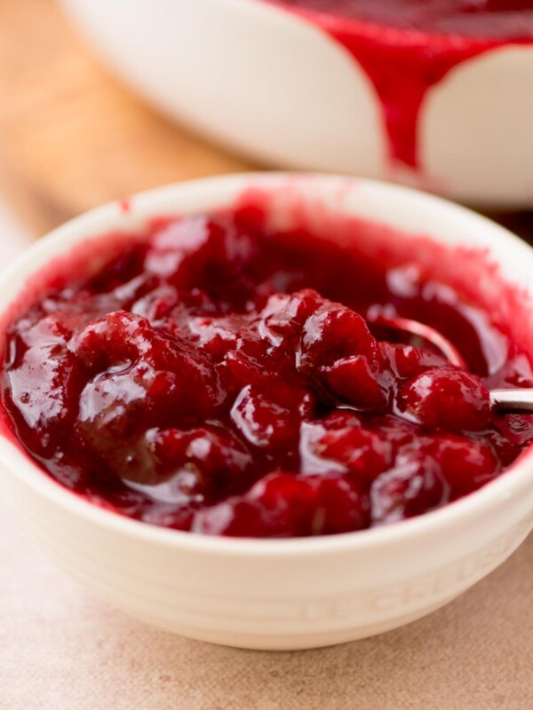 A white bowl filled with vibrant red cranberry sauce, containing whole cranberries and a glossy texture. A spoon is partially submerged in the sauce. The background shows more sauce in a white dish.