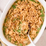 A casserole dish filled with green bean casserole topped with crispy fried onions. A spoon is scooping out a portion, revealing creamy green beans beneath the golden brown topping.