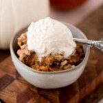 A bowl of apple crisp topped with a scoop of vanilla ice cream. The dessert is served in a gray bowl placed on a wooden surface, with a spoon resting inside the bowl. The crisp appears golden and crumbly.