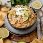 A bowl of white chicken chili garnished with lime slices and cilantro, topped with sour cream. Surrounded by tortilla chips, lime halves, and a spoon.