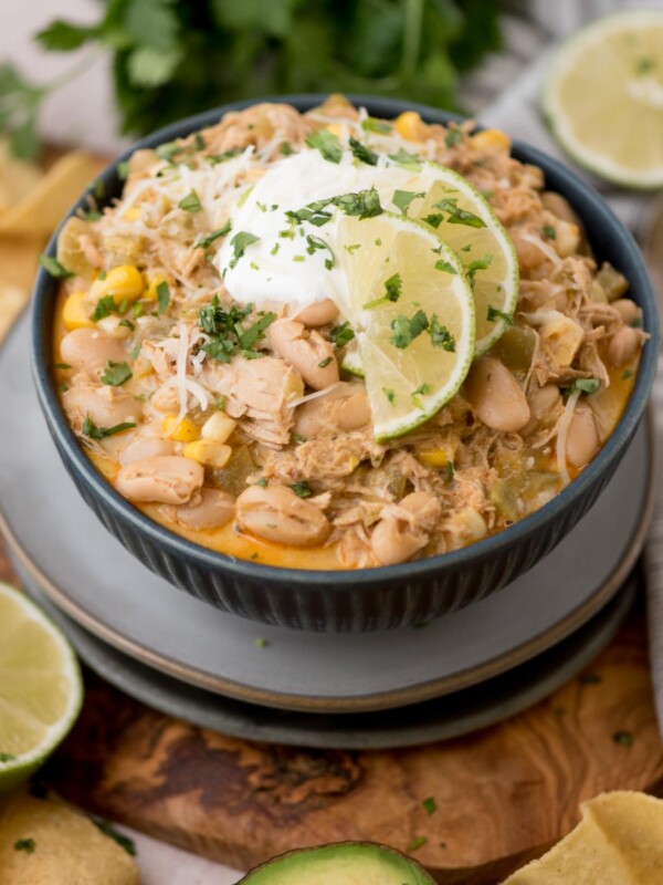 A bowl of white chicken chili garnished with lime slices and cilantro, topped with sour cream. Surrounded by tortilla chips, lime halves, and a spoon.