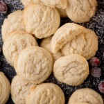 A pile of the best sugar cookies rests on a dark surface, dusted with powdered sugar. Some are broken open, revealing their soft texture. Red cranberries are scattered around these delightful Amish sugar cookies.