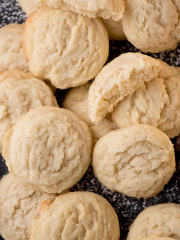 A pile of the best sugar cookies rests on a dark surface, dusted with powdered sugar. Some are broken open, revealing their soft texture. Red cranberries are scattered around these delightful Amish sugar cookies.