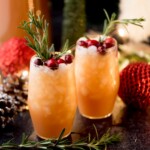 Two festive glasses of orange-colored drinks garnished with cranberries and rosemary sprigs. The background features Christmas decorations, including red ornaments and pine cones, creating a holiday atmosphere.