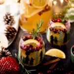 Festive holiday drinks in decorated glasses, garnished with cranberries, rosemary, and orange slices. A large pitcher filled with similar drink in the background. Pine cones, cinnamon sticks, and holiday decorations surround the setup.