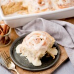 A close-up of a cinnamon roll with creamy icing on a dark plate, placed on a wooden board. A small glass with cinnamon sticks and a gray napkin sit nearby. A tray of more cinnamon rolls is in the background. A fork rests beside the plate.