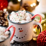 A festive mug with a snowman design is filled with hot chocolate, topped with whipped cream, colorful sprinkles, and marshmallows. Two candy canes adorn the side. In the background are holiday decorations and warm lights.