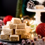 A festive stack of creamy, eggnog fudge pieces is displayed on a plate surrounded by sugared cranberries. The scene is decorated with red ornaments, pinecones, and soft fairy lights, creating a warm holiday atmosphere.