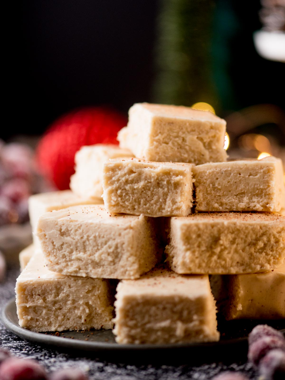 A stack of creamy white eggnog fudge pieces is piled on a plate. The fudge has a smooth texture and a dusting of nutmeg on top. Soft lighting and blurred Christmas decorations create a festive ambiance in the background.