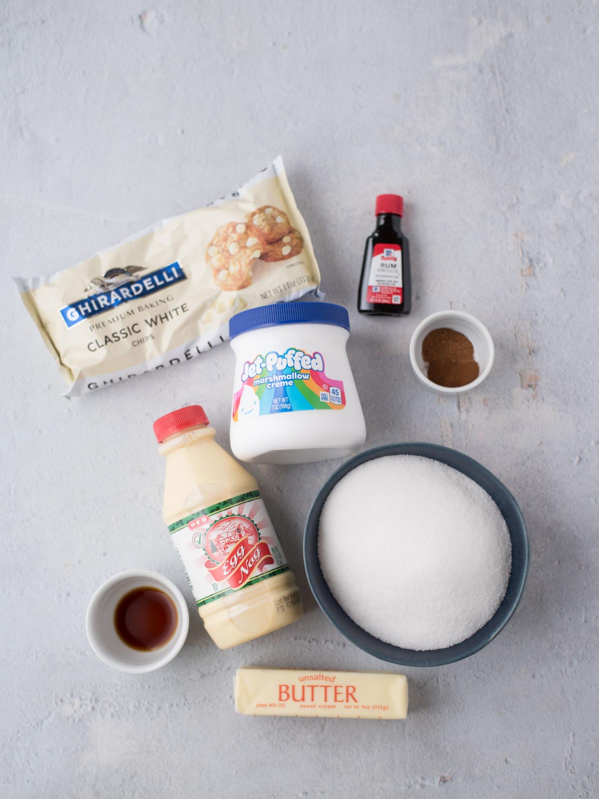 Ingredients for a festive eggnog fudge recipe are laid out on a gray surface: white chocolate chips, red food coloring, marshmallow creme, granulated sugar, evaporated milk, vanilla extract, cinnamon, and a stick of unsalted butter. .