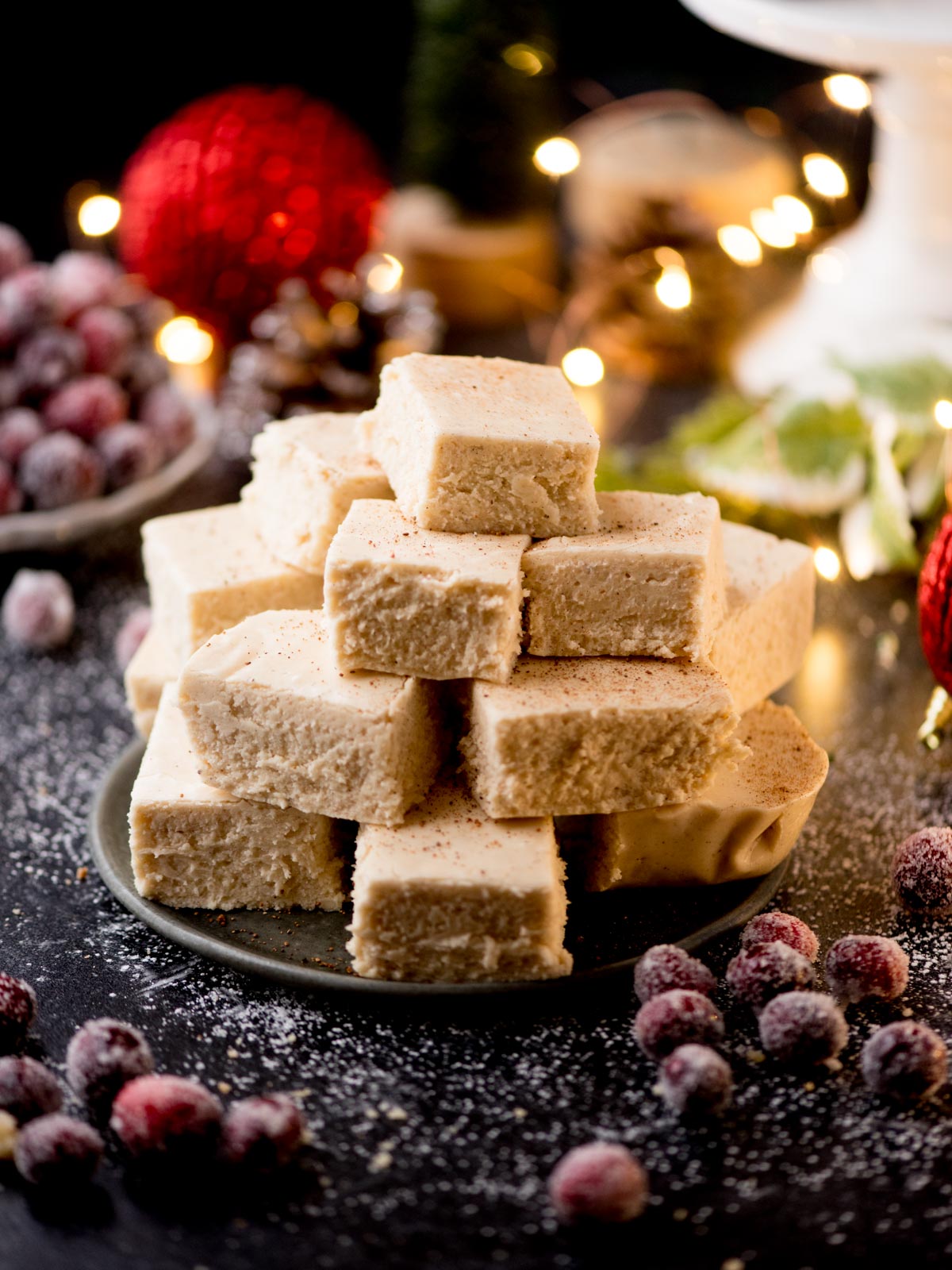 A stack of frosted cake squares sits on a plate alongside sugared cranberries, festive holiday decorations, and creamy eggnog fudge. Warm, glowing lights are blurred in the background. The scene is set on a dark surface lightly dusted with sugar.